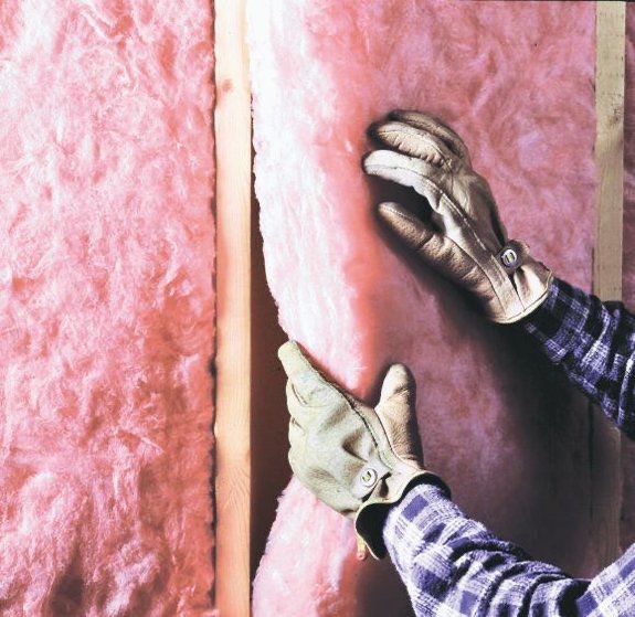 Gloved hands pressing a fiberglass batt into place between wall studs
